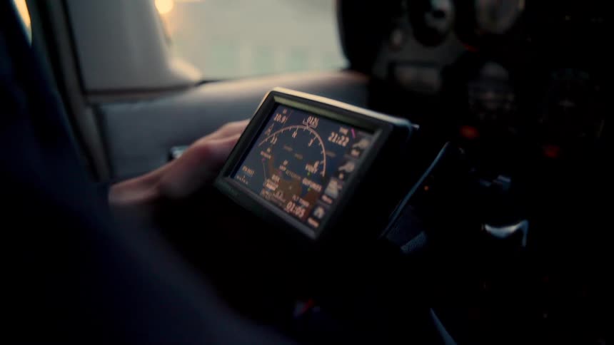A pilot operates a handheld flight computer while taxiing a small aircraft past a lit hangar