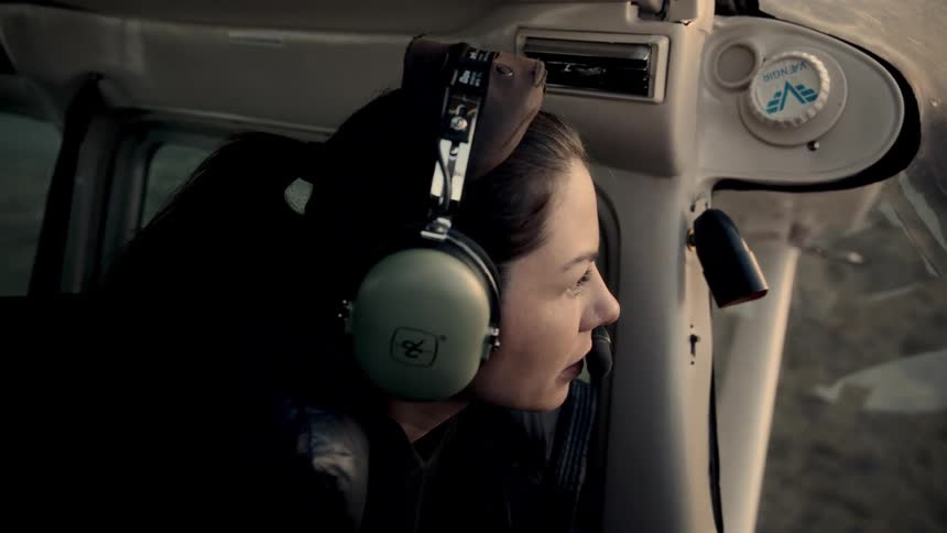 A woman wearing large green headphones and a microphone sits in the cockpit of a small airplane and looks out the side window at the landscape below during a flight at sunset