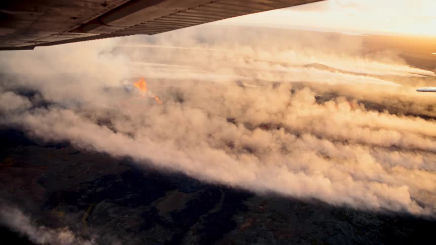 A forest fire sweeps across an open field at sunset sending thick smoke upward until it spreads through the sky and clouds the atmosphere