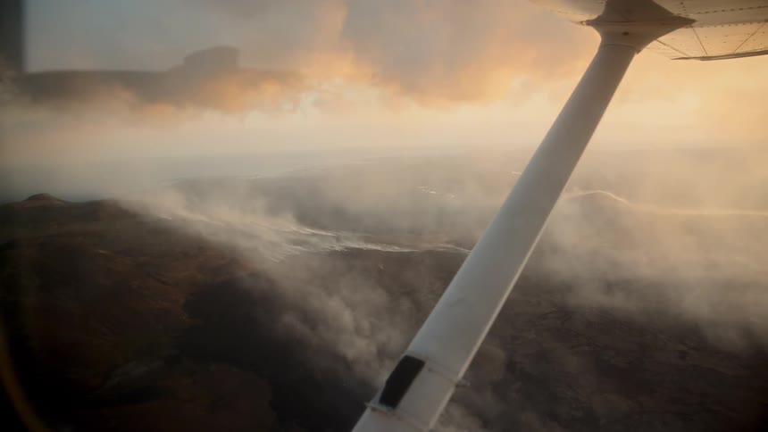 A view from an airplane window shows a wide landscape covered in thick white smoke under a golden sky at sunset