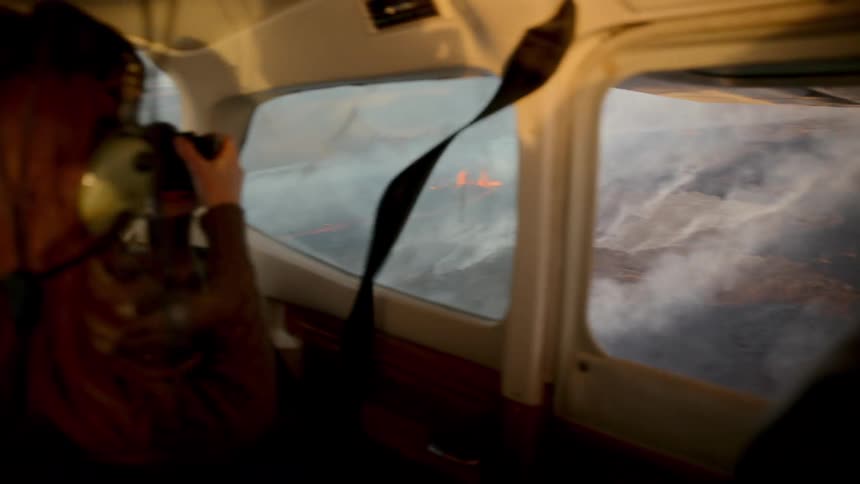 A person sitting inside a small airplane takes photos through a side window at a volcanic eruption with orange lava and thick smoke on the ground below