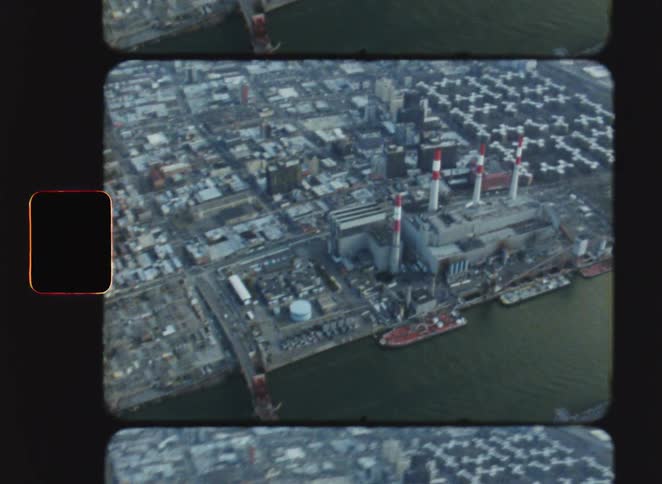 The aerial footage shows a vintage birdseye view of a coastal city prominently featuring a power plant with tall redandwhite striped smokestacks as the camera pans it reveals the industrial waterfront and a large bridge stretching across the river
