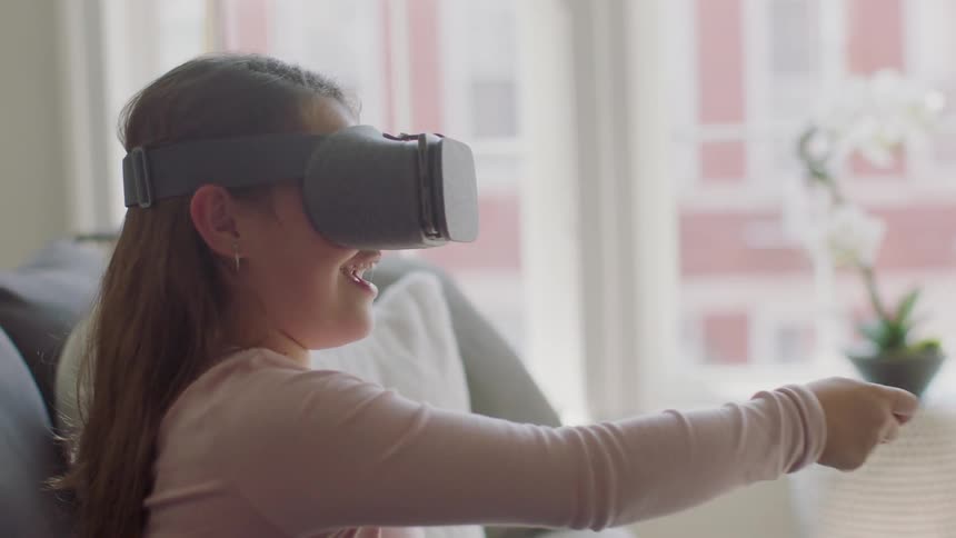 Little girl sitting on couch wearing virtual reality headset and playing game