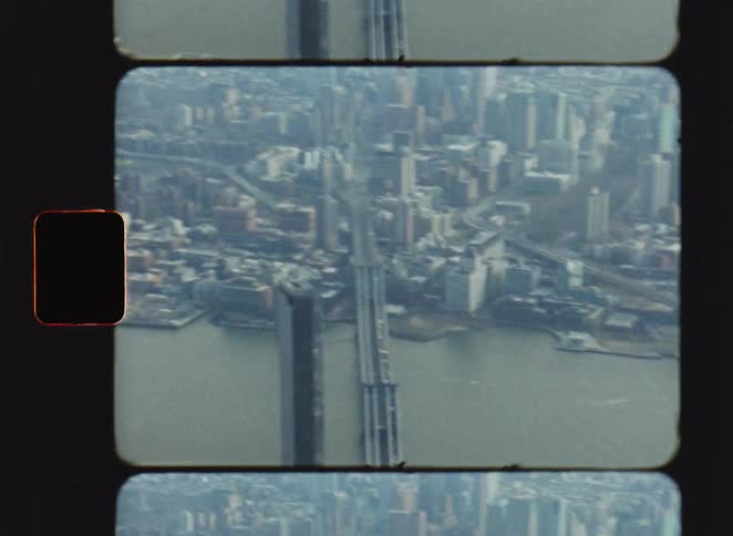 Vintage film footage captured on an eight millimeter camera shows a high view of a long bridge crossing over a river in a city with many buildings and a green field visible on the shore under a hazy sky