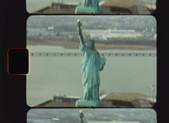 Vintage film footage shows a high view of the statue of liberty standing on liberty island with the surrounding water and distant industrial shore visible under a gray sky