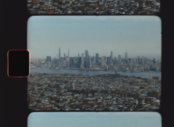 An aerial shot of a city skyline shows several skyscrapers and a river while the camera moves slowly over residential rooftops in the foreground