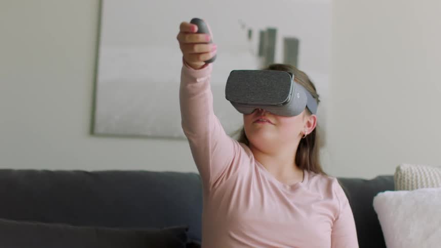 Young girl sitting on a couch with a controller in her hand held out in front of her while wearing virtual reality headgear