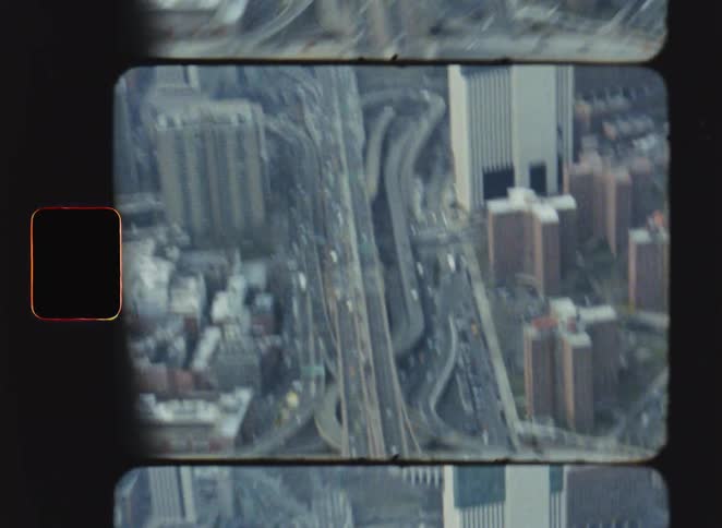 Vintage film footage shows a high view of a long bridge filled with cars crossing over a body of water in a city with highways and tall buildings visible on the shore