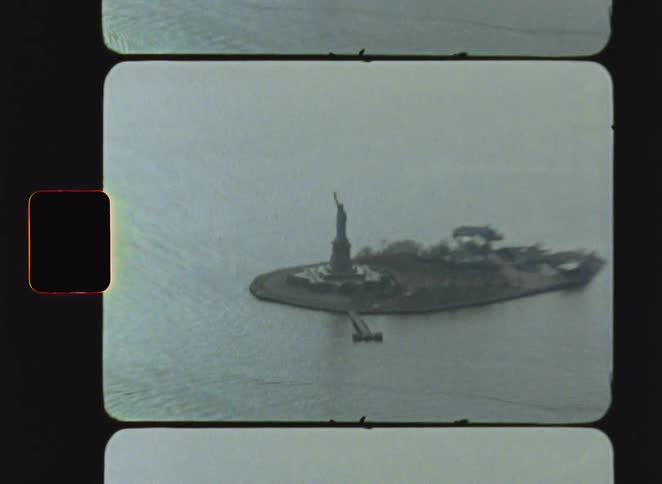 An aerial vintage film shot shows the statue of liberty standing on liberty island surrounded by the harbor water under an overcast sky