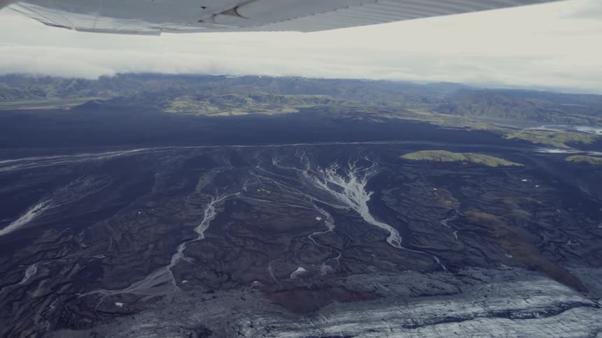 A distant view from an airplane shows a vast dark landscape of braided river channels and volcanic plains with rolling green hills and mountains under a cloudy sky