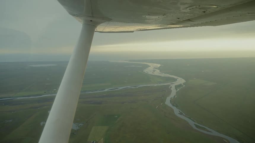 Wing section visible above flat fields crossed by winding river under overcast sky