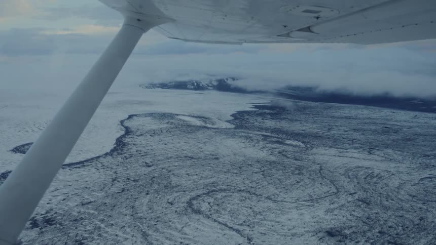 Aircraft wing and strut visible flying over glacial landscape with low clouds and mountains in the distance