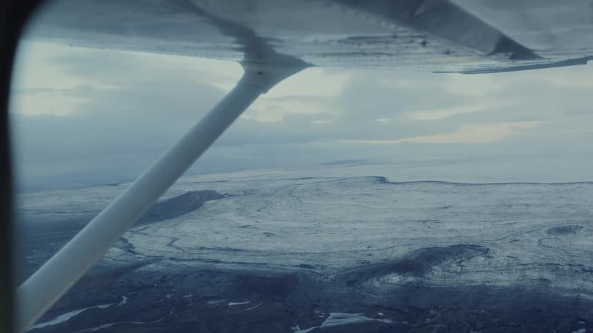 Wing and strut visible flying over icy landscape with dark streaks under overcast sky