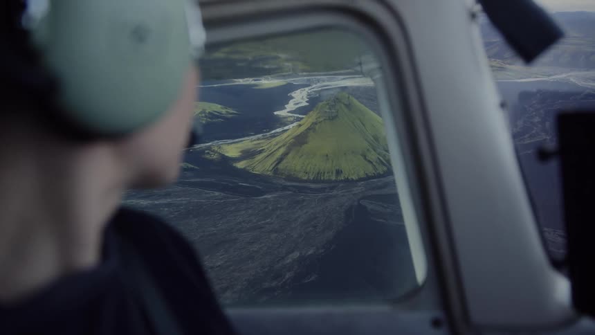 A person wearing green headphones looks out of an airplane window at a bright green volcanic mountain surrounded by dark lava fields and winding rivers