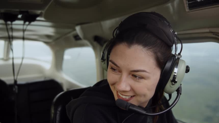 Adult young woman wearing a pilot headset on the cabin of an aircraft smiling and talking to people around her indoors during day time