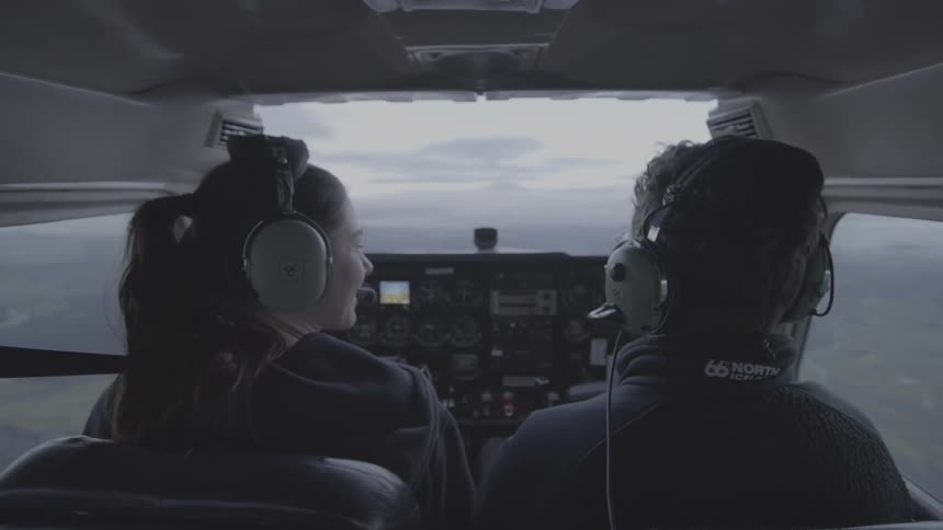 A view from behind two people in the cockpit of a small airplane shows them looking ahead at the cloudy horizon during a flight
