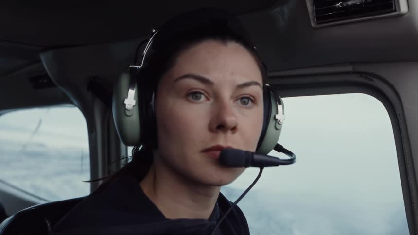 A woman wearing green headphones sits in the cockpit of a small airplane and looks out the side window during a flight over a snowy landscape