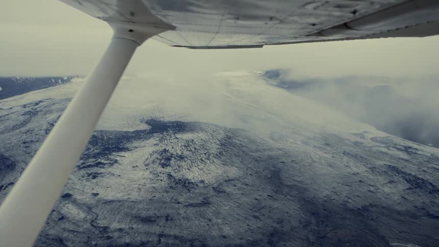 An aerial view from an airplane shows a vast and dark glacial landscape covered in patches of white snow under a thick layer of grey clouds
