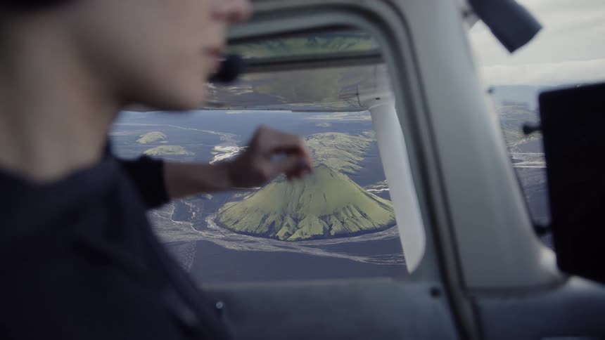 Young adult female pilot on an airplane opening the cabin window towards the view of a mountain range indoors during day time