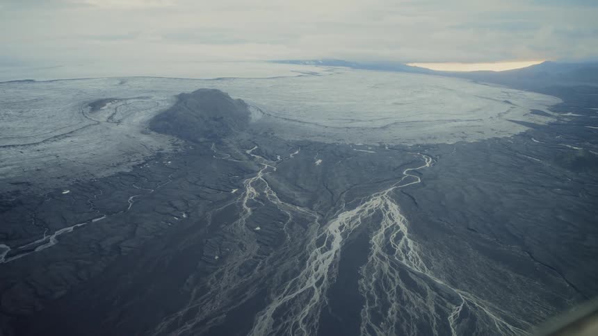 A high view from a plane shows a vast white glacier meeting a dark landscape of braided river channels under a cloudy sky
