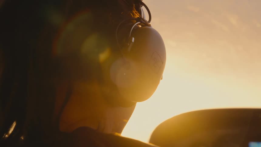 A woman wearing a headset is silhouetted against the brilliant golden light of a setting sun inside an airplane cockpit