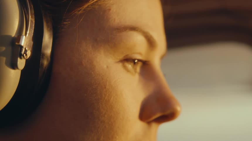 A close up shot shows the face of a woman wearing large headphones as she looks forward during a flight at sunset
