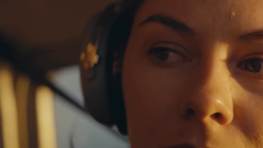 A close view of a woman pilot wearing a headset inside a cockpit looks ahead and slightly to the side while warm golden sunlight shines on her face during a flight at sunset