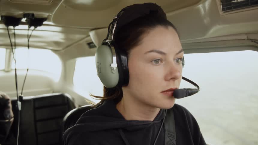 A young woman pilots a small airplane at midday sunlight pouring through the cockpit windows shes wearing a flight helmet focused on the controls while a passenger sits in the seat behind her