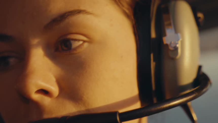 A close view of a woman pilot wearing a headset inside a cockpit looks ahead and slightly to the side while warm golden sunlight shines on her face during a flight at sunset