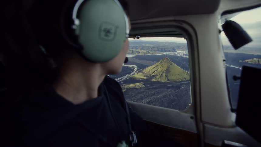 A young woman sits in the passenger seat of an airplane wearing headphones as she lifts her phone to snap a photo of the mountains below while the plane flies overhead