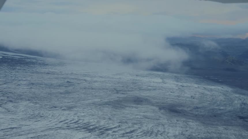 Aircraft wing flying over glacier with volcanic terrain in background under overcast sky and distant light