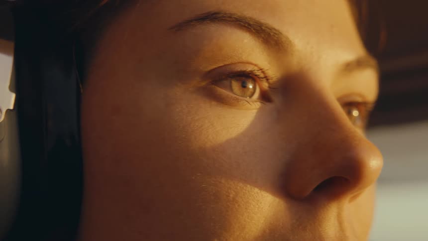A close view of a woman pilot wearing a headset inside a cockpit looks ahead and slightly to the side while warm golden sunlight shines on her face during a flight at sunset