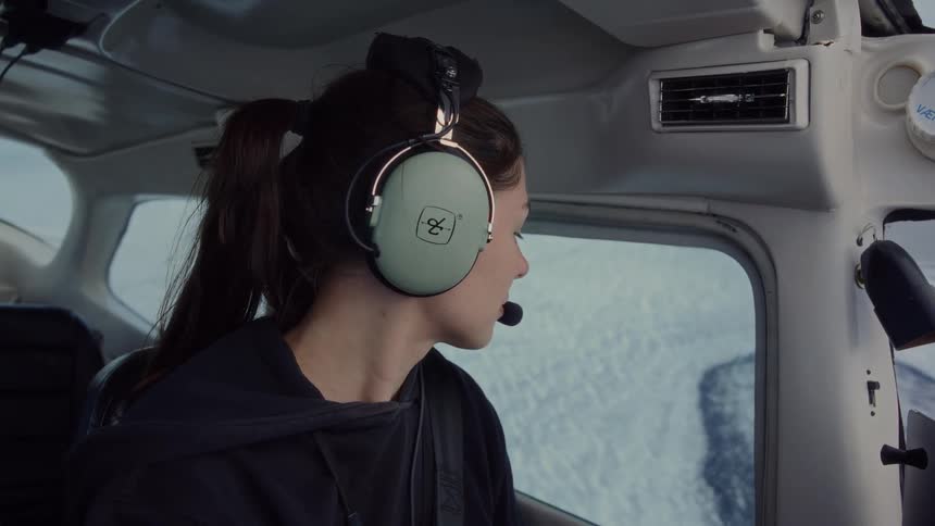 A young woman wearing a black hoodie and large green headphones sits inside a small airplane and looks out the window at a snowy landscape below