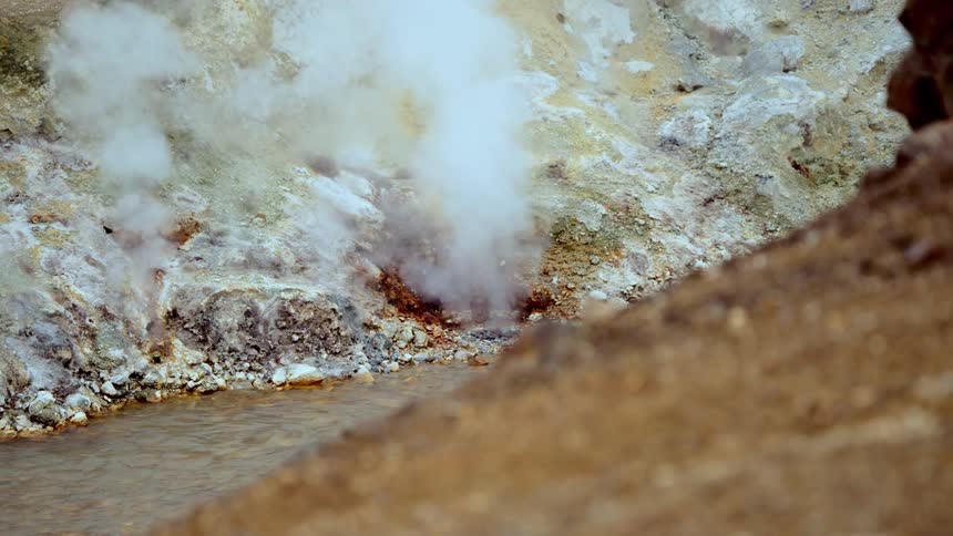 Smoke rises from hot lava beside a flowing stream at midday the bright light catching the steam as it lifts into the air and mixes with the heat shimmering above the rocks