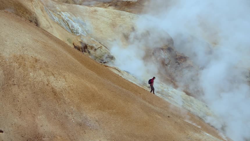 A lone hiker carefully descends a steep steam shrouded volcanic mountain in a rugged geothermal landscape