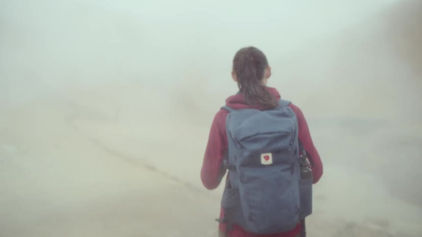 A view from behind of a woman with a ponytail wearing a red jacket and a blue backpack as she walks through thick white steam in a geothermal area with brown hills in the distance