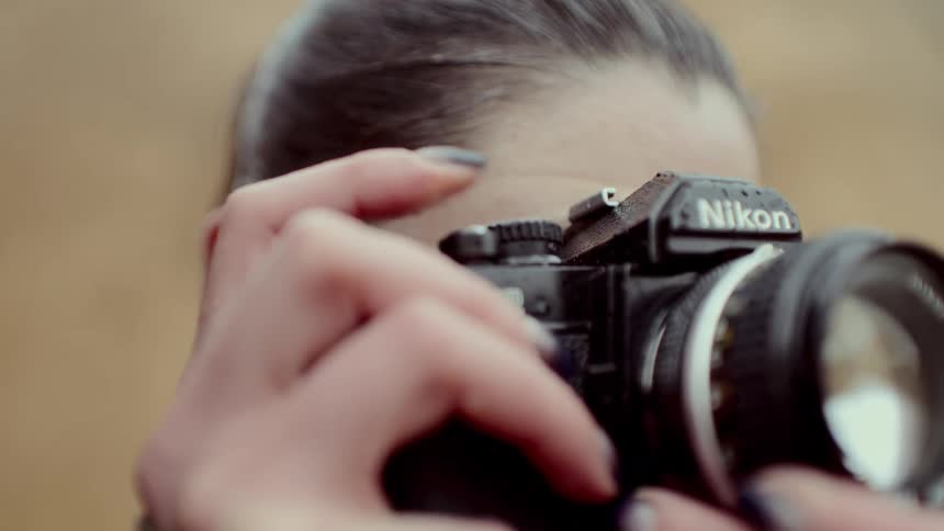 A close view of a woman with her hair in a ponytail as she raises a black vintage film camera to her eye to take a photo outdoors