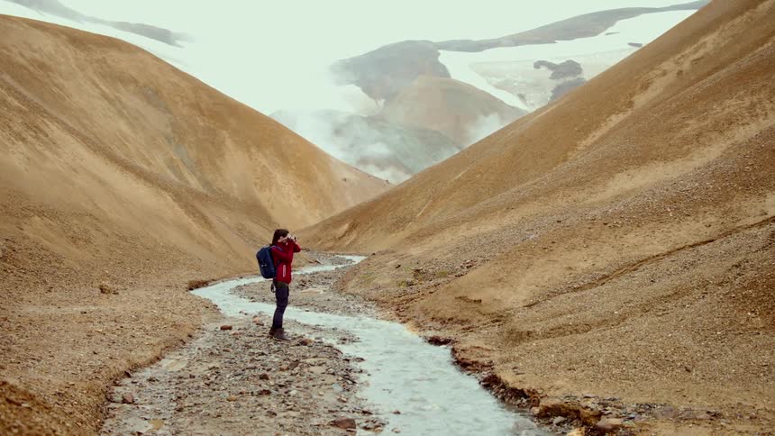 A person wearing a red jacket and a blue backpack stands by a stream in a valley and takes photos of the tan hills and snowy mountains