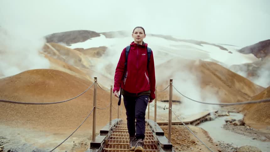 Woman walks along a walkway in a geothermal area with steam and mountains in the background