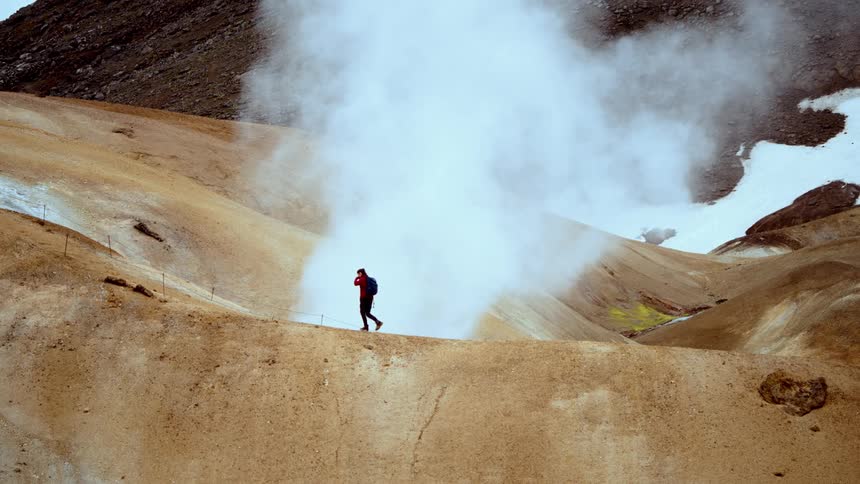 A person hikes on a volcanic mountain at midday carrying a backpack as smoke rises from the mountains surface