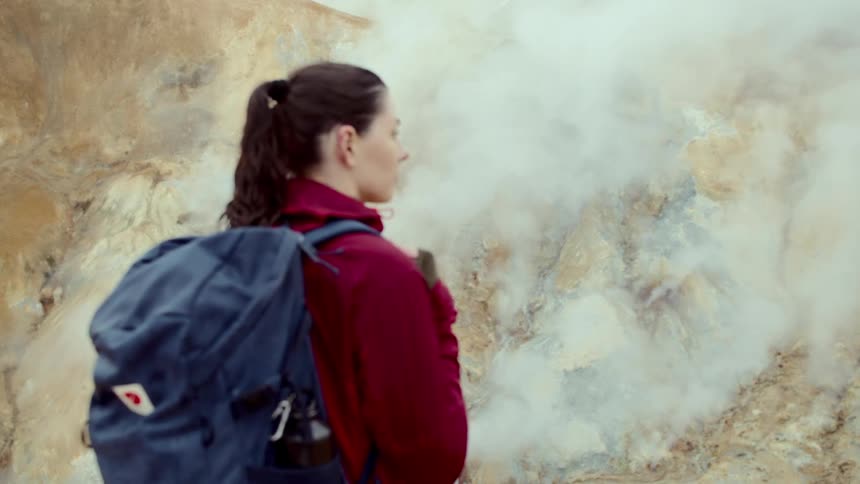 A young woman with a backpack hikes through the wild at midday sunlight bright on the terrain she pauses on the trail as she notices smoke rising from a patch of burning grass in the distance