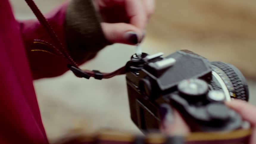A close view of a woman wearing a red jacket as she uses her fingers to carefully thread a thin black strap through the metal eyelet of a vintage film camera outdoors