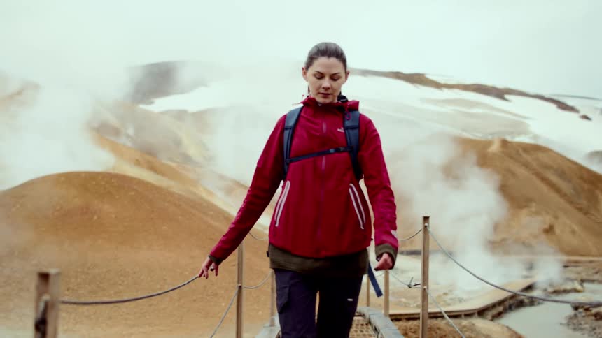 A woman wearing a red jacket and a blue backpack walks across a wooden bridge in a geothermal landscape with steam rising from brown hills and snow in the distance