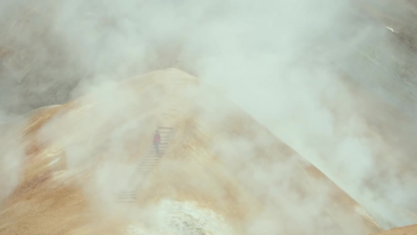 A distant view shows a person wearing a red jacket walking down a set of wooden stairs on a tan hillside while thick white steam from geothermal vents drifts across the landscape