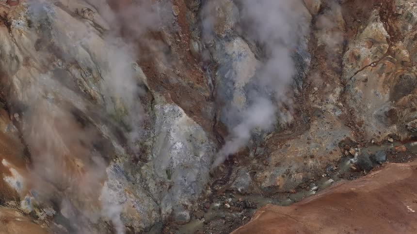 A high view of a rocky geothermal area shows thick white steam rising from various vents in the brown and gray earth
