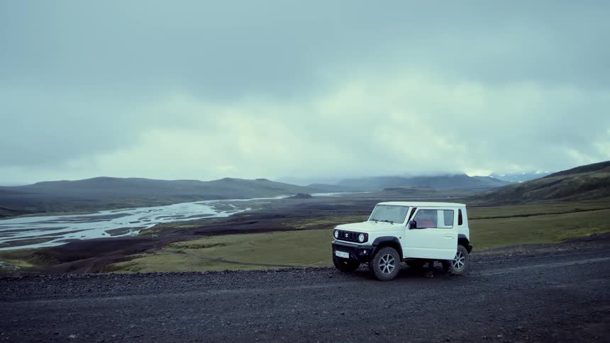 A woman wearing a red jacket and a blue backpack walks along a gravel road toward a white off road vehicle parked in a vast mountain landscape with a winding river under a cloudy sky