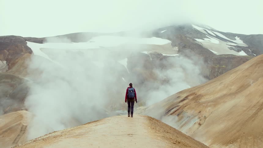A woman with a blue backpack and red jacket walks along a narrow dirt path toward a geothermal area with steam rising from vents in a rocky mountain landscape under a cloudy sky