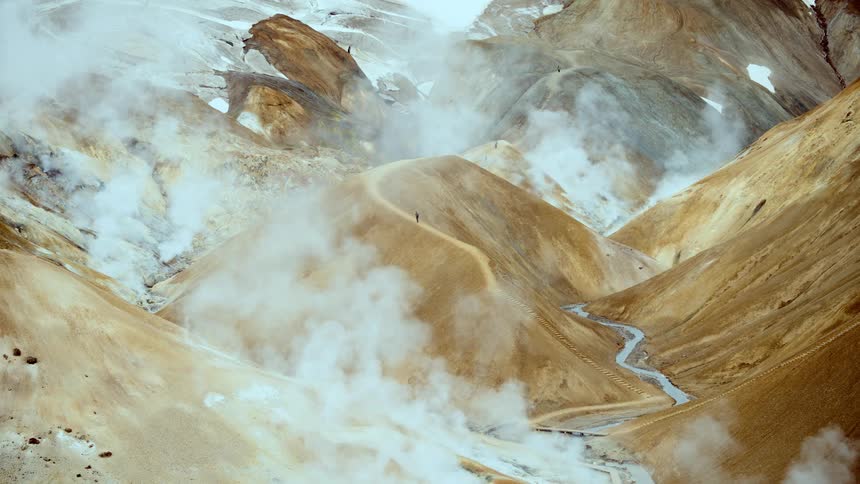 A distant view of a person walking along a narrow ridge in a vast geothermal mountain landscape with thick white steam rising from the valley and a small blue stream flowing below
