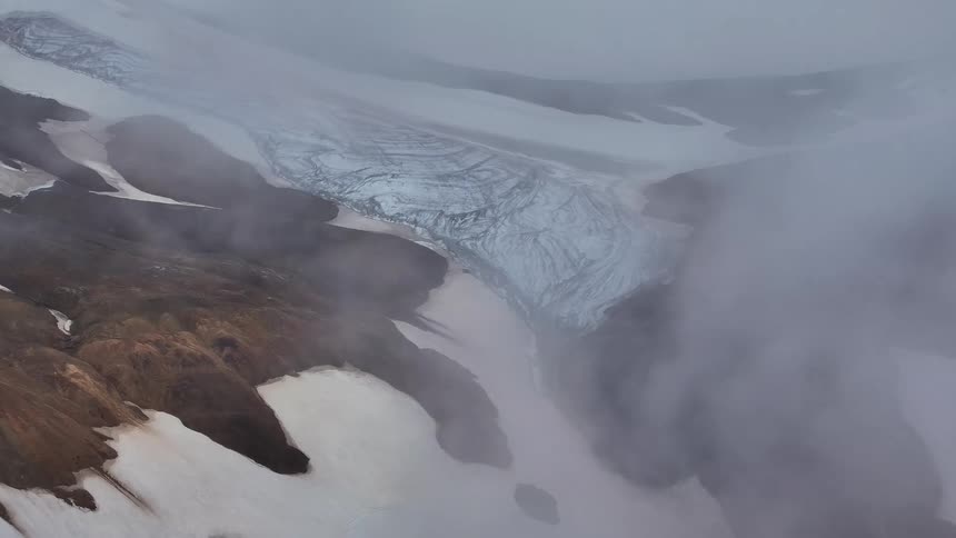 Blue glacier winds its way between rocky ridges under mist in a mountainous region