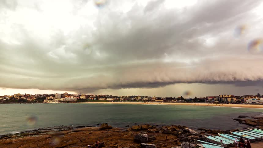 A distant view of a beach and coastal city under a vast shelf cloud as a storm moves in and rain begins to fall on the lens